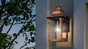 The Urban Ambiance UQL1413 Vintage Outdoor Wall Light from the Longview Collection, featuring a rustic copper finish and classic gaslight-inspired design, is mounted on a modern house exterior, casting a warm glow at dusk with tree branches visible to the left.