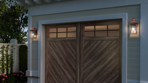 A wooden garage door with a chevron pattern is flanked by two Urban Ambiance UQL1413 Vintage Outdoor Wall Lights from the Longview Collection, featuring a rustic copper finish, on a light blue house exterior.