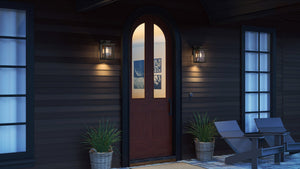 A wooden arched door with frosted glass is framed by Urban Ambiance UQL1642 Craftsman Outdoor Wall Sconces, potted plants, and outdoor chairs on a porch with dark siding.
