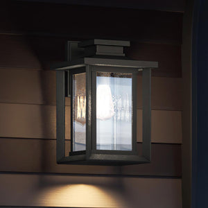 An Urban Ambiance UQL1643 Craftsman Outdoor Wall Sconce from the Peacehaven Collection, with a natural black finish and marine-grade construction, illuminates a dark brown paneled wall at dusk.