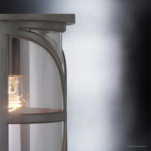 Close-up of the Urban Ambiance UHP1182 Rustic Outdoor Pendant Light from the Brussels Collection, featuring a 22″ x 7-1/2″ die-cast aluminum frame in an Olde Bronze finish, with a lit bulb shining through its curved glass enclosure, set against a gradient gray background.
