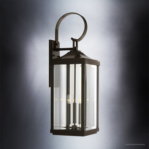 The Urban Ambiance UHP1194 Colonial Outdoor Wall Light from the Calderdale Collection features an Olde Bronze finish with a rectangular beveled glass enclosure and three visible candle-style bulbs, beautifully illuminated against a gray background.